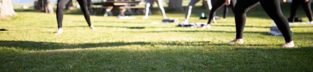 6 yoga practitioners stand in a Warrior II, barefoot in the grass at Dancing Spirit Ranch in Montanan. There is a picnic table in the background, water bottles and yoga mats nearby.